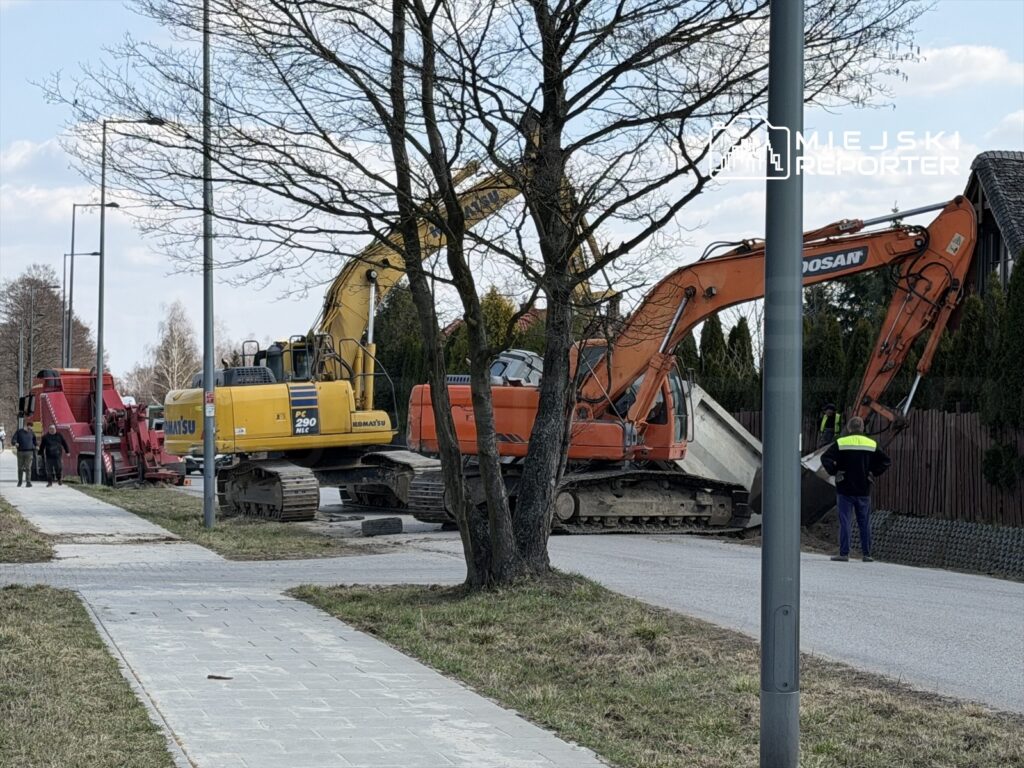 Trzy maszyny budowlane, w tym żółty koparka i pomarańczowy sprzęt, pracują przy modernizacji drogi w otoczeniu drzew.