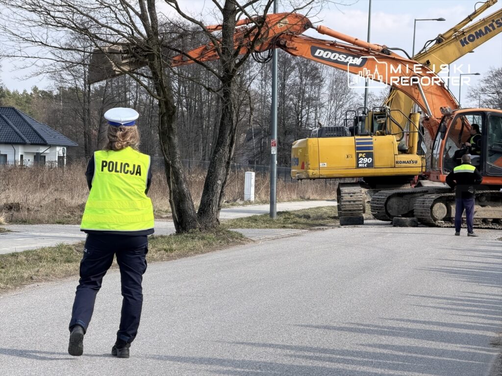 Policjantka w kamizelce odblaskowej obserwuje pracę maszyny budowlanej na ulicy w otoczeniu drzew i domów.