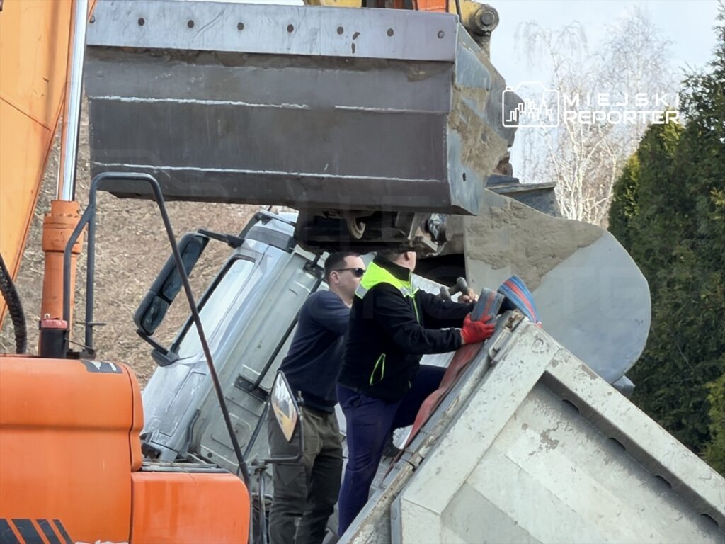 Dwaj mężczyźni usiłują podnieść przewrócony pojazd ciężarowy przy użyciu koparki w terenie leśnym.