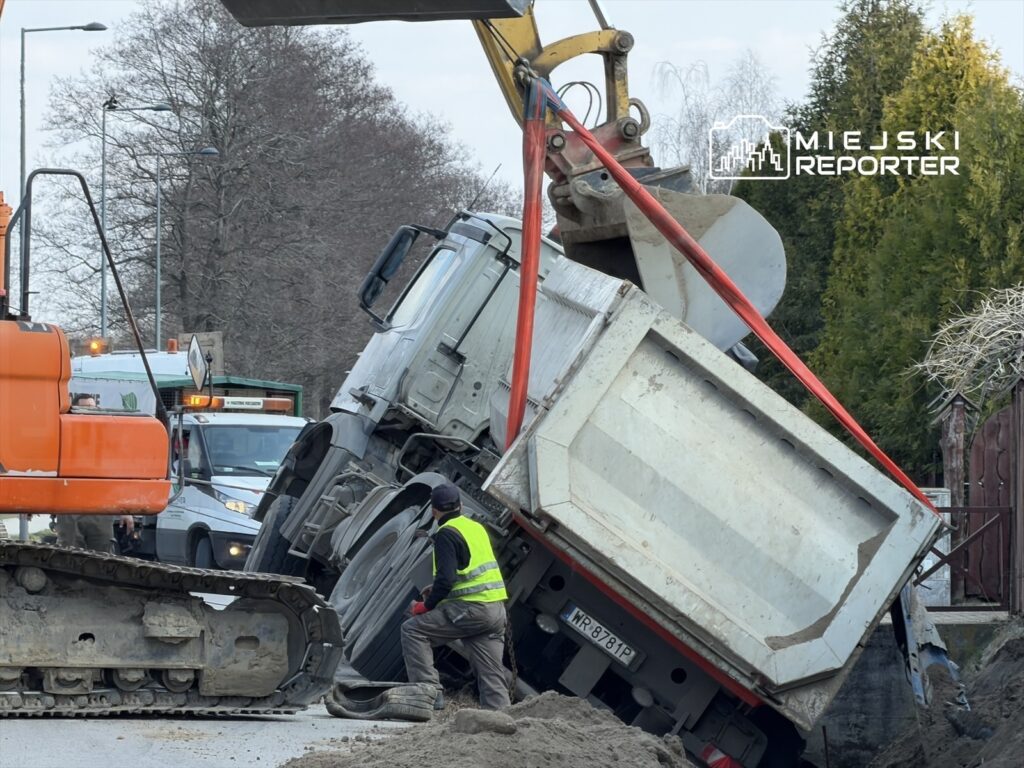 Ciężarówka z przyczepą przewrócona na boku, podczas akcji ratunkowej w miejskim otoczeniu, z użyciem koparki.