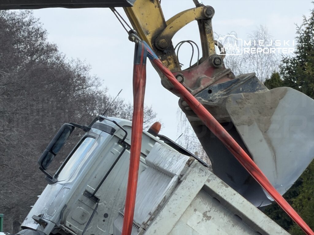 Koparka podnosi przewrócony ciężarówkę na tle drzew.