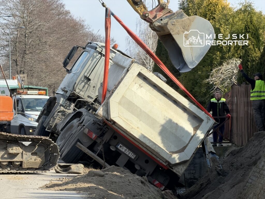 Ciężarówka z przechyloną skrzynią ładunkową jest podnoszona przez dźwig na drodze, obok pracujący robotnicy.