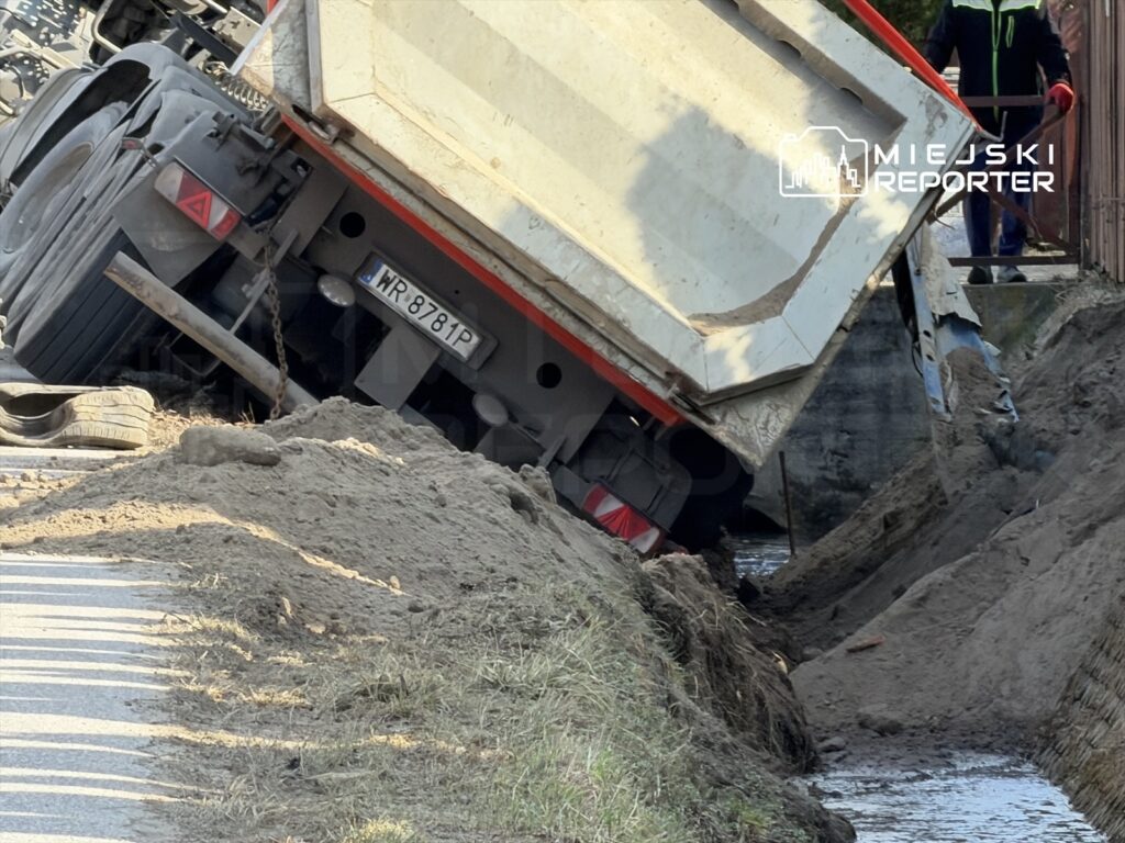 Cementowóz przewrócony na boku przy drodze, obok wykopu, z widocznymi oponami i piaskiem na ziemi.
