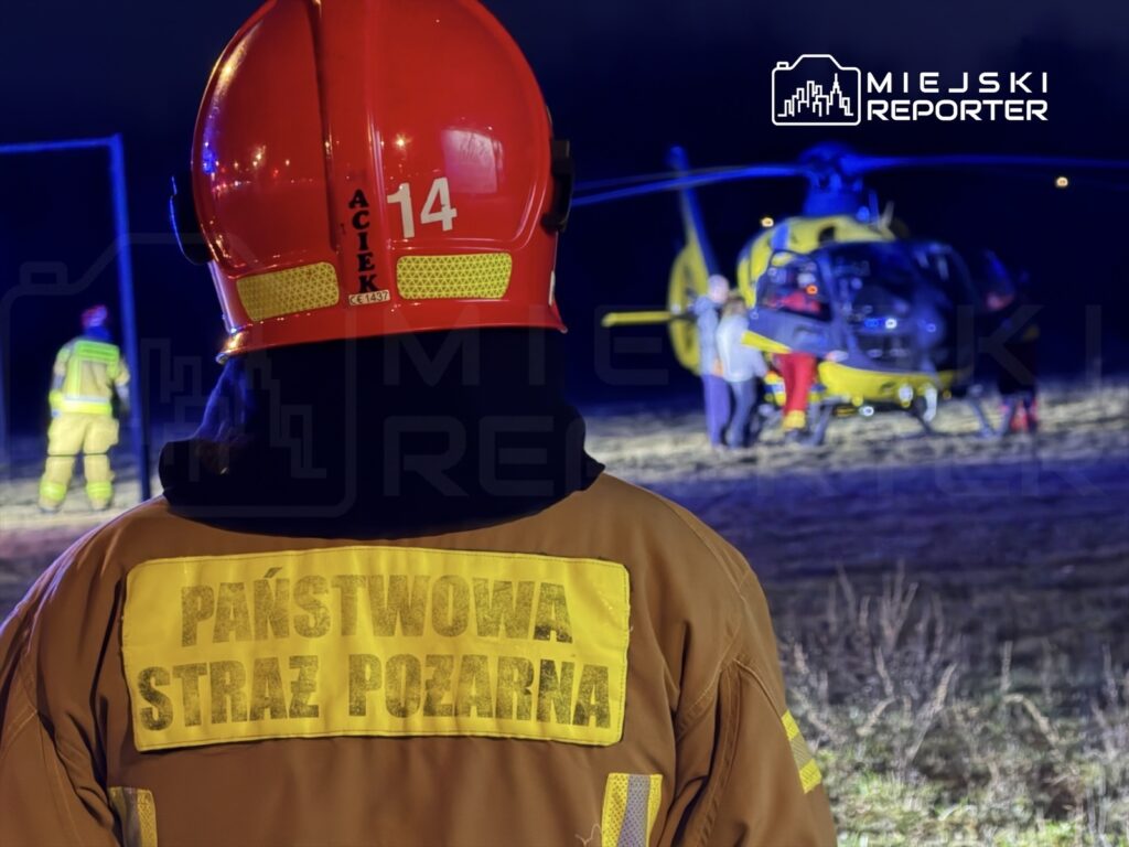 Strażak w hełmie z numerem 14 obserwuje akcję ratunkową przy śmigłowcu w nocy.