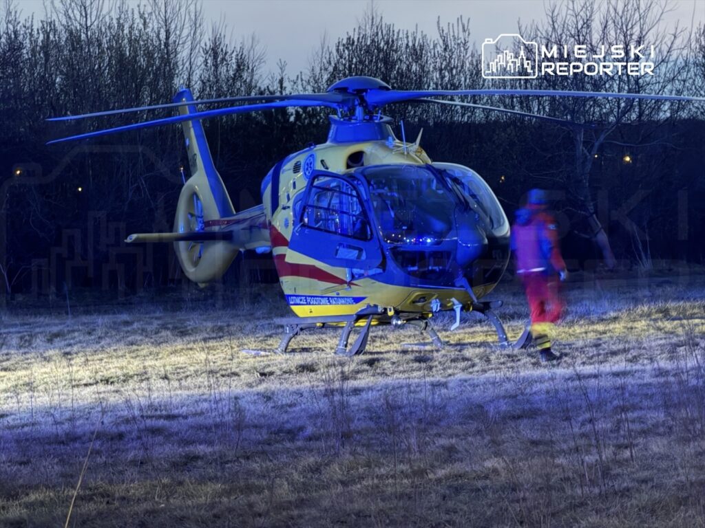 Śmigłowiec ratunkowy ląduje na trawiastym terenie, obok którego przechodzi ratownik w odzieży ochronnej.