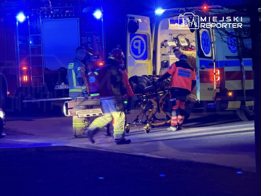 Grupa ratowników medycznych transportuje osobę na noszach do karetki w otoczeniu wozów strażackich i niebieskich świateł.