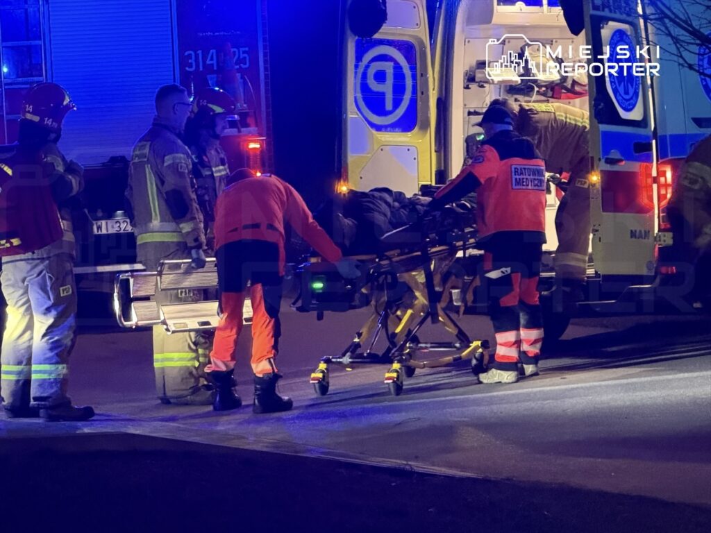 Grupa ratowników medycznych i strażaków przenosi poszkodowanego na noszach do karetki w nocy.
