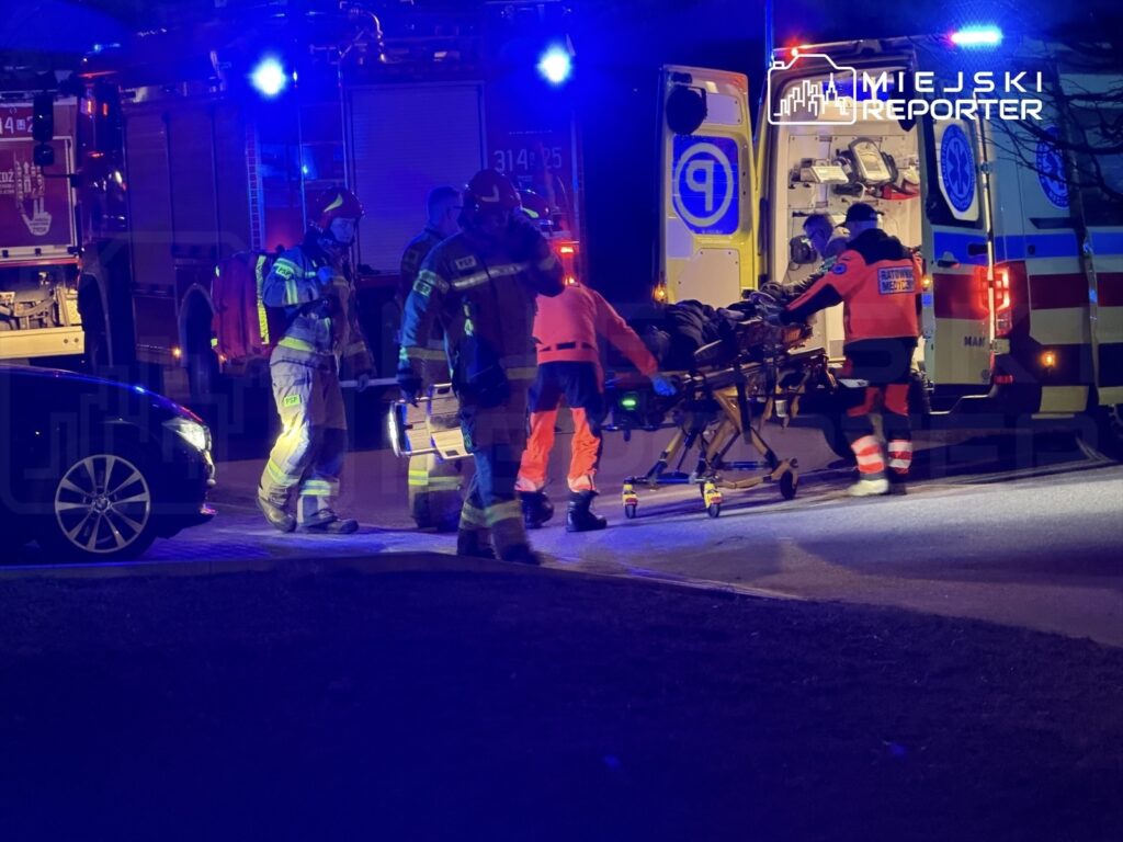 Grupa ratowników medycznych i strażaków przenosi nosze z poszkodowanym obok wozów strażackich i karetki w nocy.