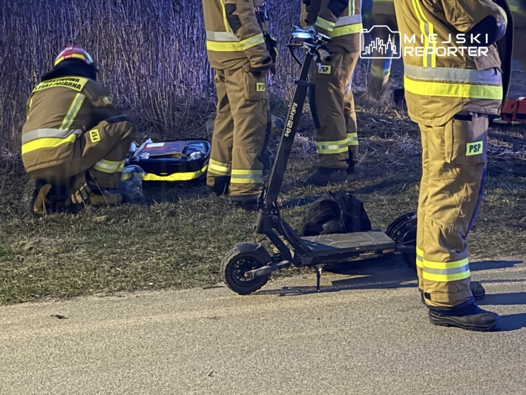 Grupa strażaków w specjalistycznych strojach pracuje obok uszkodzonego skutera elektrycznego na trawie.