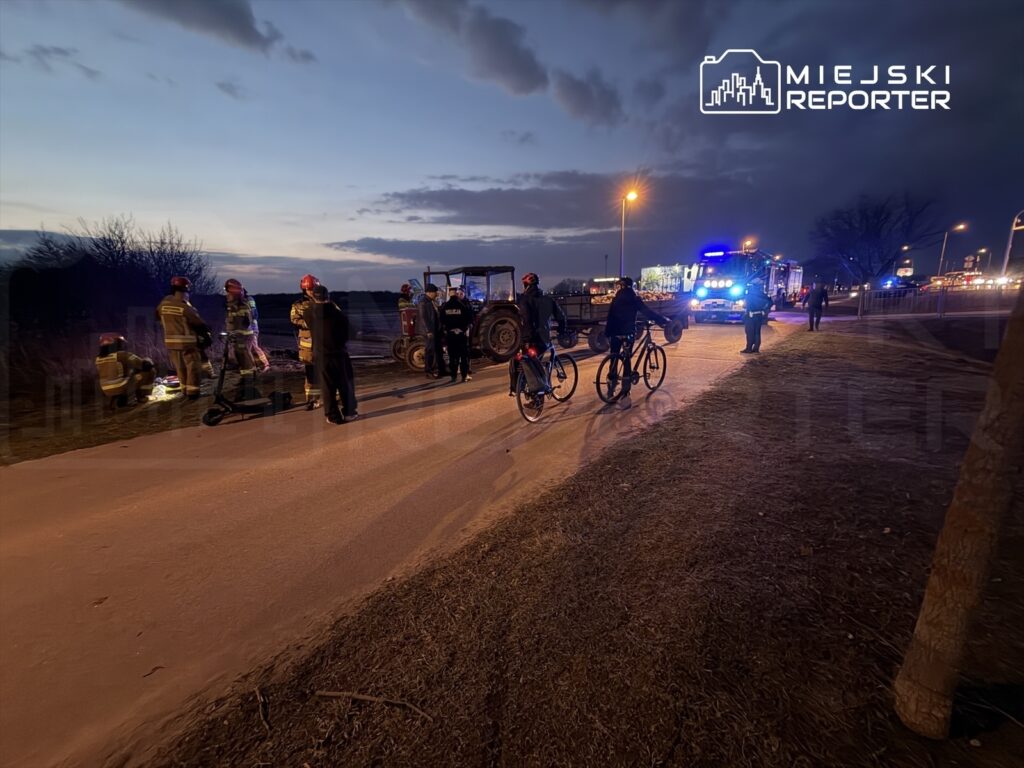 Grupa strażaków i policjantów pracuje na drodze, obok stoi traktor, a rowerzyści obserwują sytuację w zmierzchu.