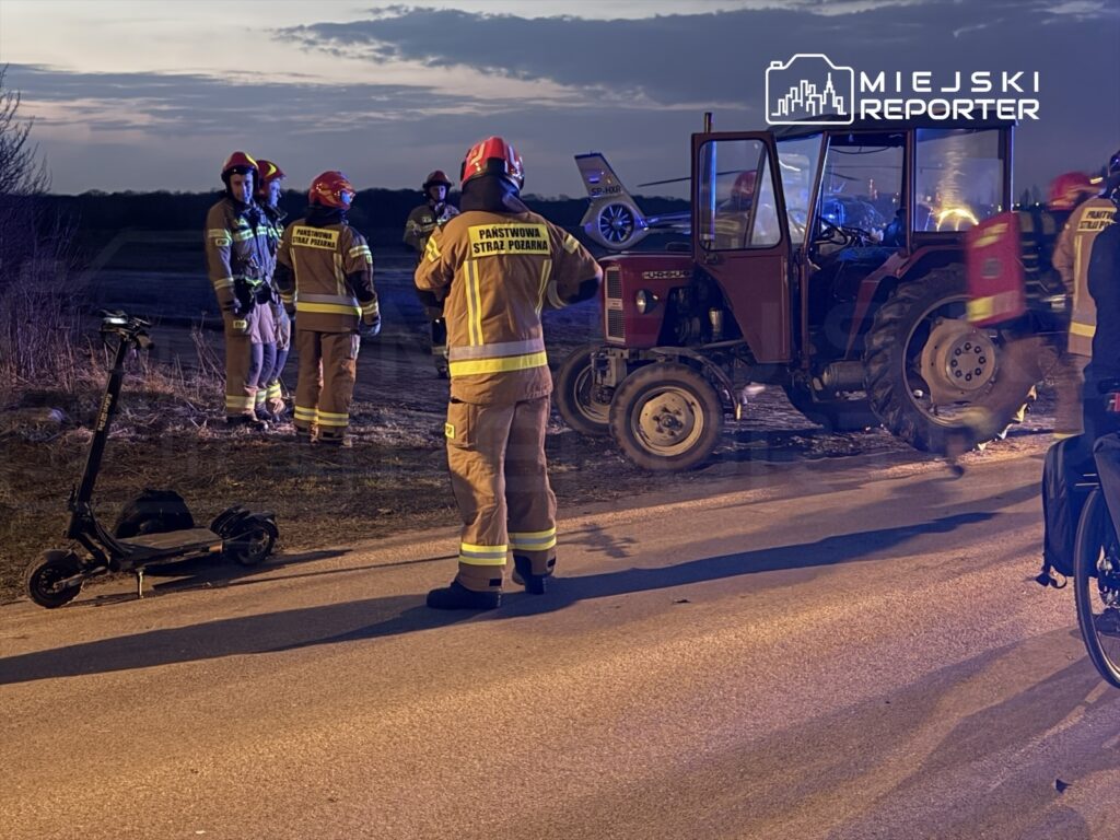 Grupa strażaków w mundurach i hełmach pracuje przy uszkodzonym ciągniku na drodze, obok leży elektryczna hulajnoga.