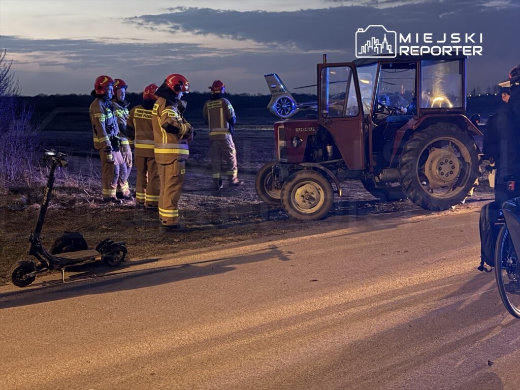 Grupa strażaków w hełmach obserwuje uszkodzony traktor na drodze, obok leży elektryczna hulajnoga.