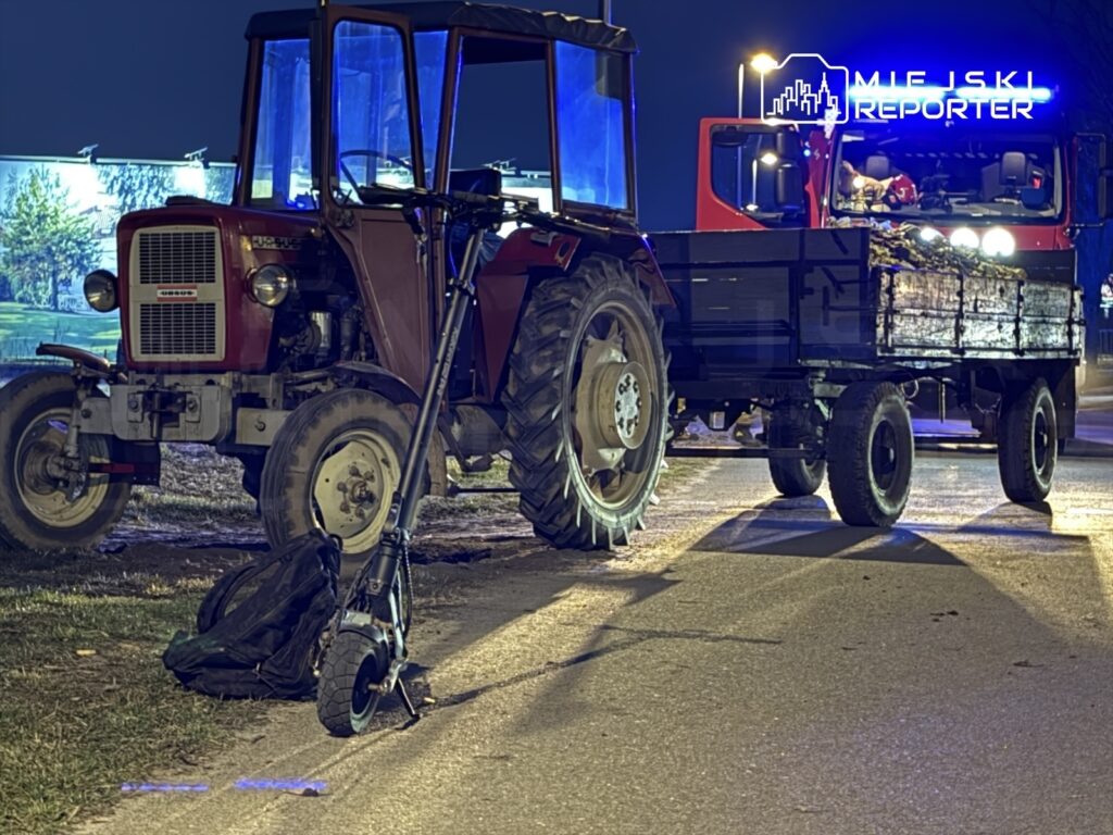 Traktor z czerwoną kabiną stoi obok ciężarówki z ciemnym przyczepą na oświetlonej ulicy w nocy, obok leży hulajnoga.