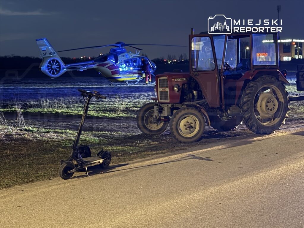 Traktor stojący przy drodze obok elektrycznej hulajnogi, w tle lądujący helikopter ratunkowy.