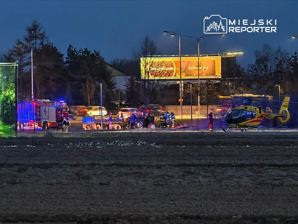 Helikopter ratunkowy ląduje w pobliżu akcji ratunkowej, otoczony przez strażaków i pojazdy na drodze.