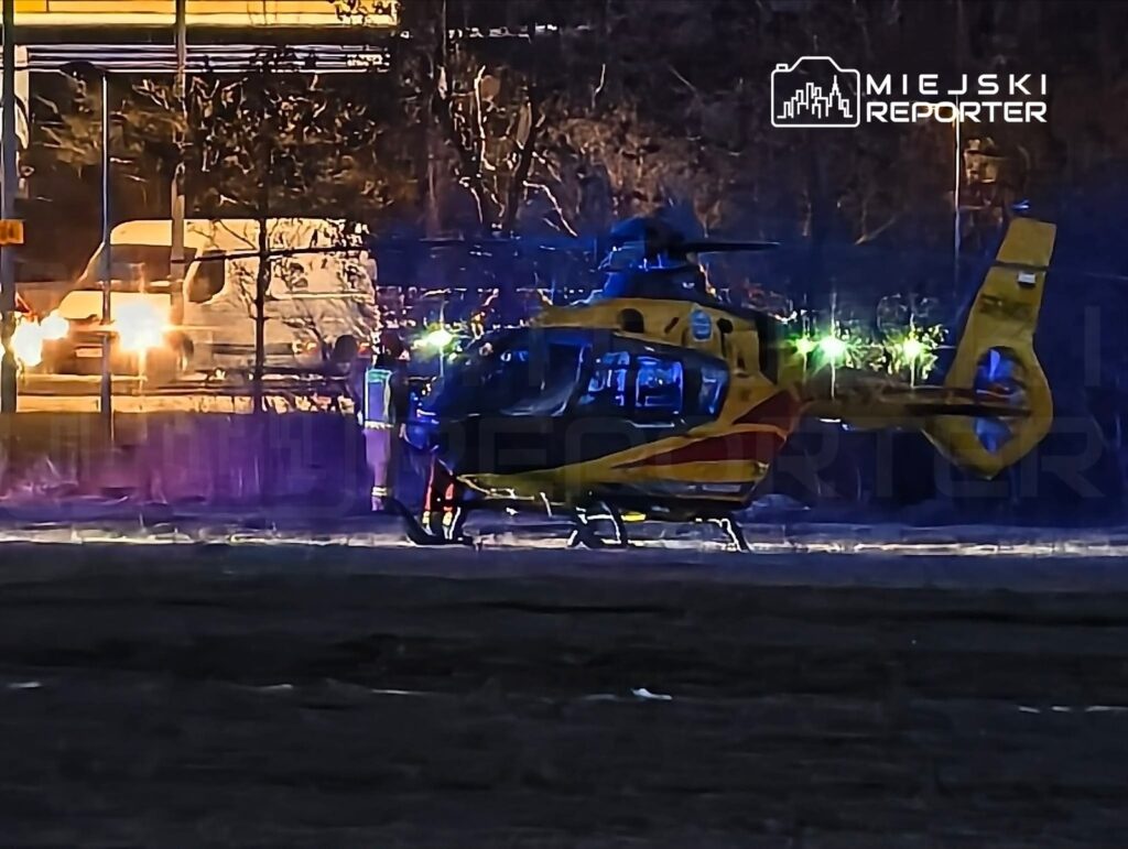 Śmigłowiec ratunkowy ląduje na nieoświetlonym terenie, w tle widać pojazdy poruszające się po drodze.