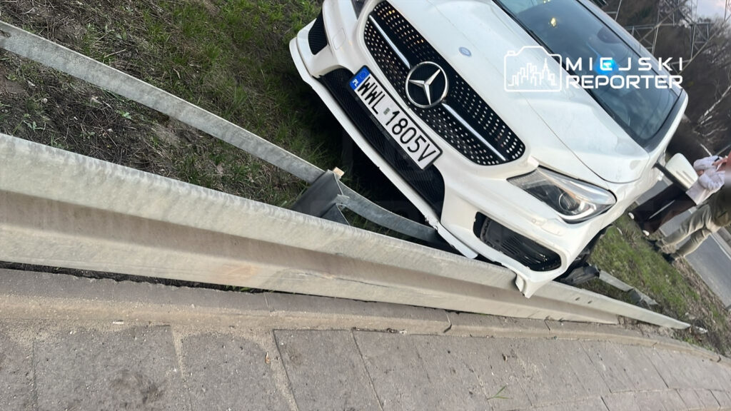 Biały samochód osobowy marki Mercedes stoi na poboczu, zniszczona bariera ochronna leży obok. W tle widać przechodniów.