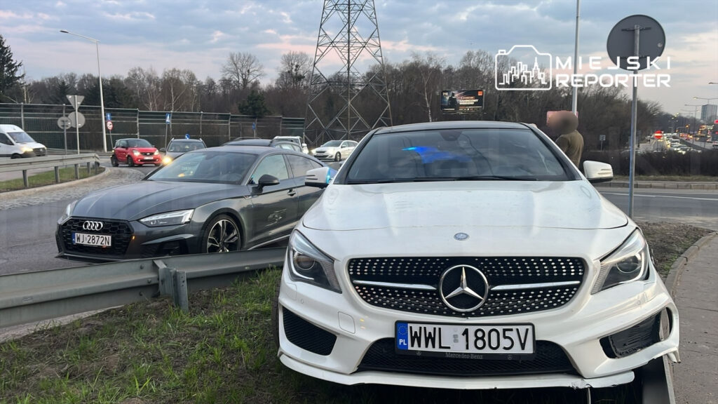 Dwa samochody osobowe, biały Mercedes oraz szary Audi, stoją na poboczu drogi w miejskim otoczeniu. W tle widać ruch uliczny oraz billboard.