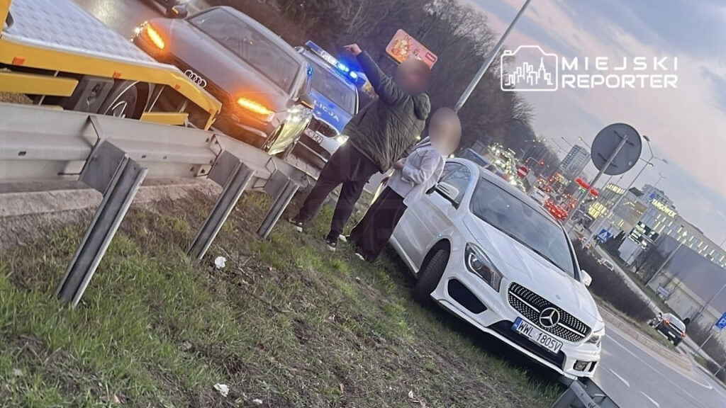 Mężczyzna w ciemnej kurtce wskazuje na policyjny radiowóz obok białego samochodu osobowego na poboczu drogi.