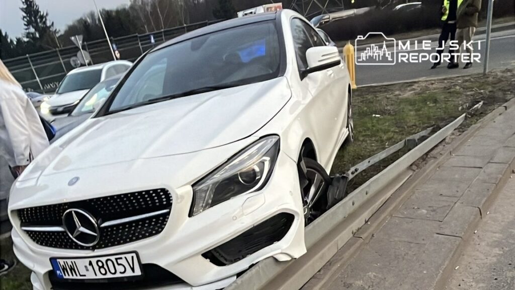 Biały samochód osobowy marki Mercedes stoi na poboczu drogi, z uszkodzoną felgą i unieruchomionym kołem. W tle widać inne pojazdy oraz ludzi w odzieży roboczej.