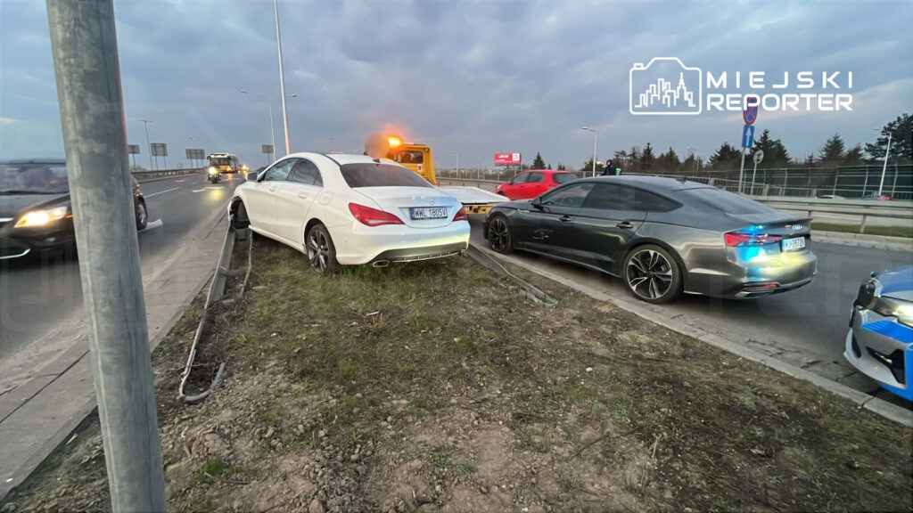 Biały samochód osobowy utknął na poboczu drogi, obok niego stoi szary pojazd, a w tle widać inne samochody na jezdni.