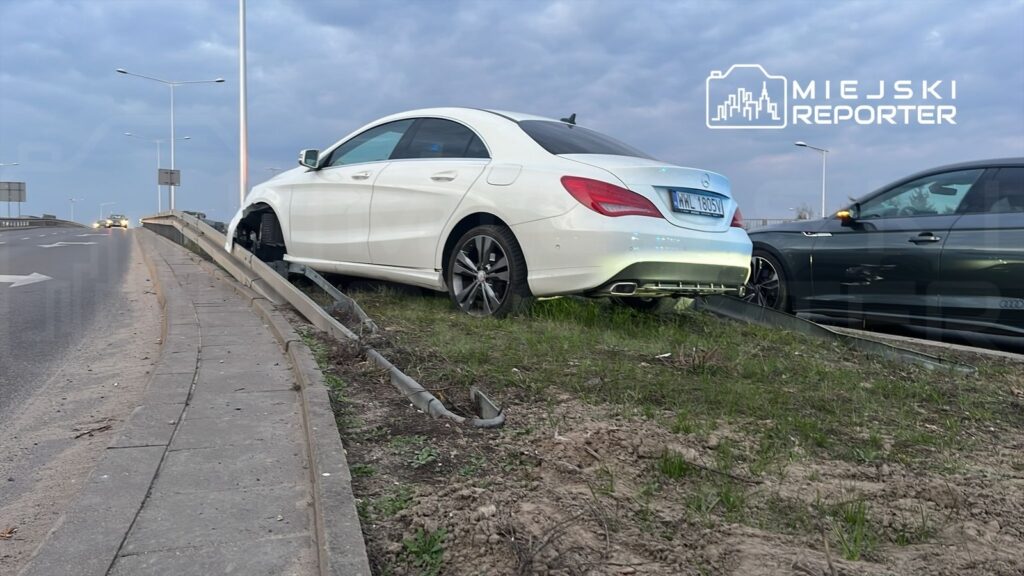 Biały samochód osobowy zaparkowany na trawie obok drogi, z uszkodzoną przednią osią, obok ciemny pojazd.