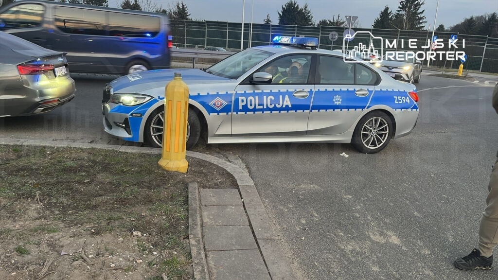 Policyjny samochód osobowy z niebieskimi światłami zatrzymany przy krawężniku, obok żółtej barierki ochronnej.