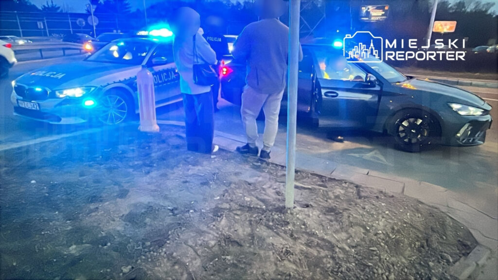 Policjant rozmawia z dwoma osobami obok radiowozu z włączonymi niebieskimi światłami na drodze.