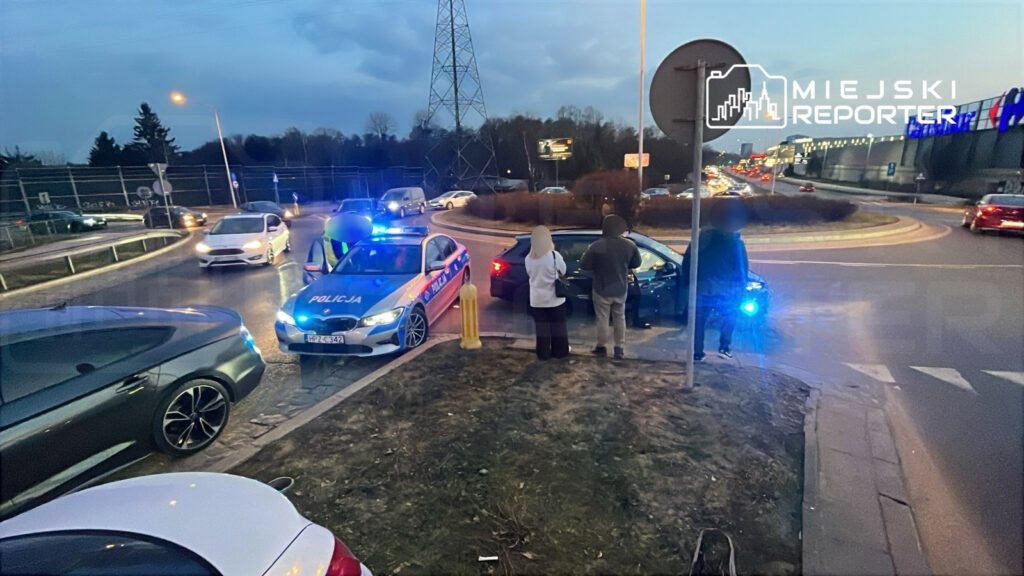 Policja kontroluje pojazd na rondzie, w tle widoczne inne samochody i billboardy.