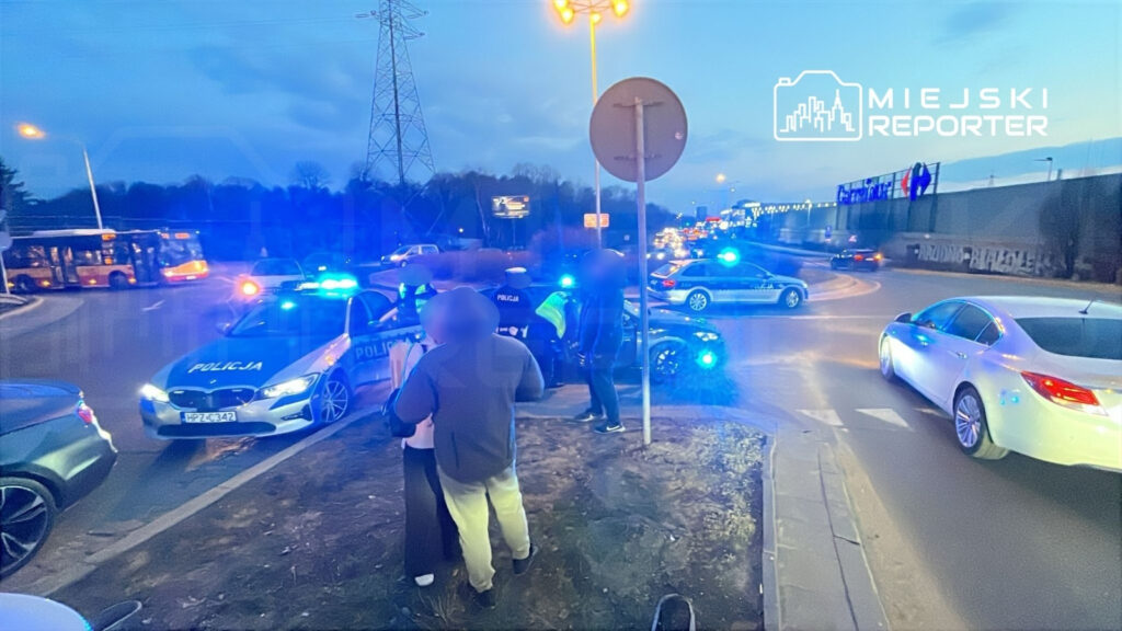 Grupa osób rozmawia z policjantami obok radiowozów z włączonymi sygnałami świetlnymi na skrzyżowaniu.