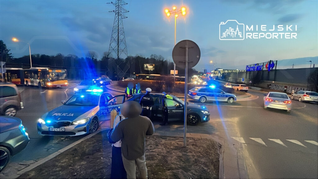 Policjanci z włączonymi sygnalizatorami zatrzymują ruch na skrzyżowaniu, a przechodnie obserwują sytuację.
