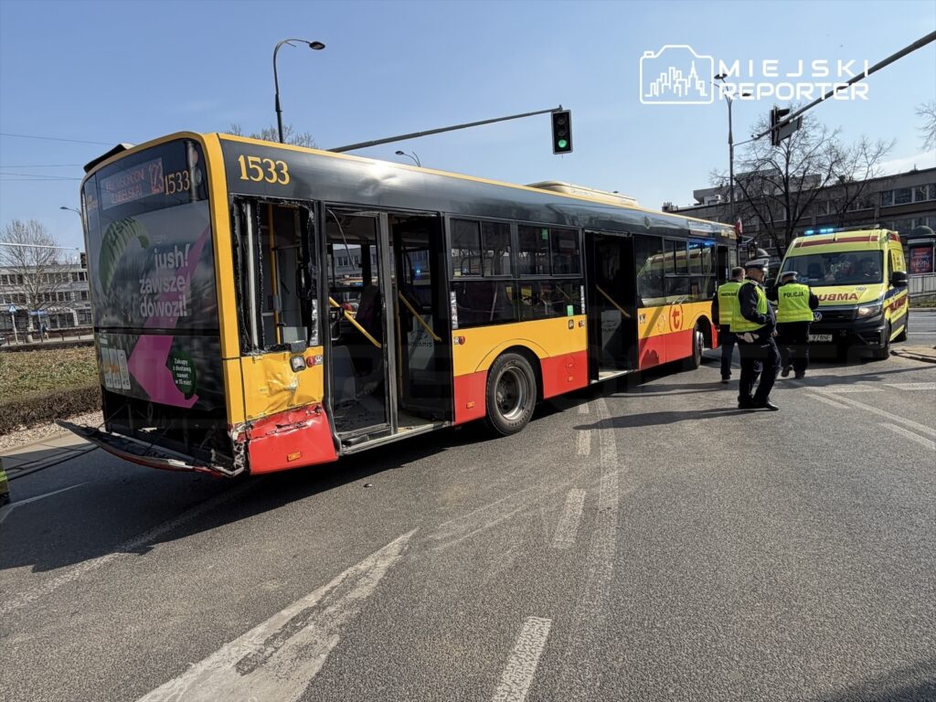Autobus z uszkodzeniem tyłu stoi na skrzyżowaniu, a obok niego pracują policjanci i zespół ratunkowy.