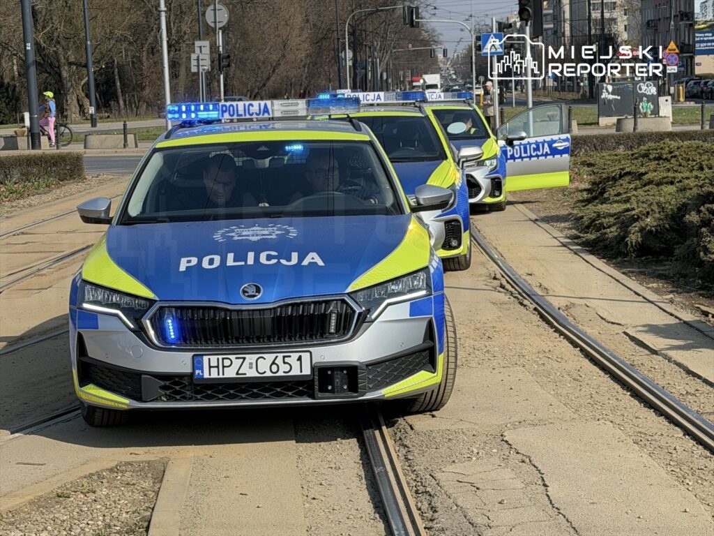 Policjanci w radiowozie z niebieskimi światłami zatrzymani na torowisku w miejskim otoczeniu.