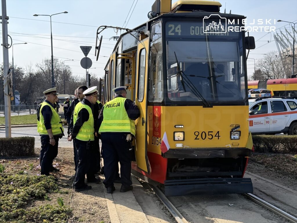 Grupa policjantów w kamizelkach odblaskowych rozmawia przy tramwaju linii 24 na przystanku.