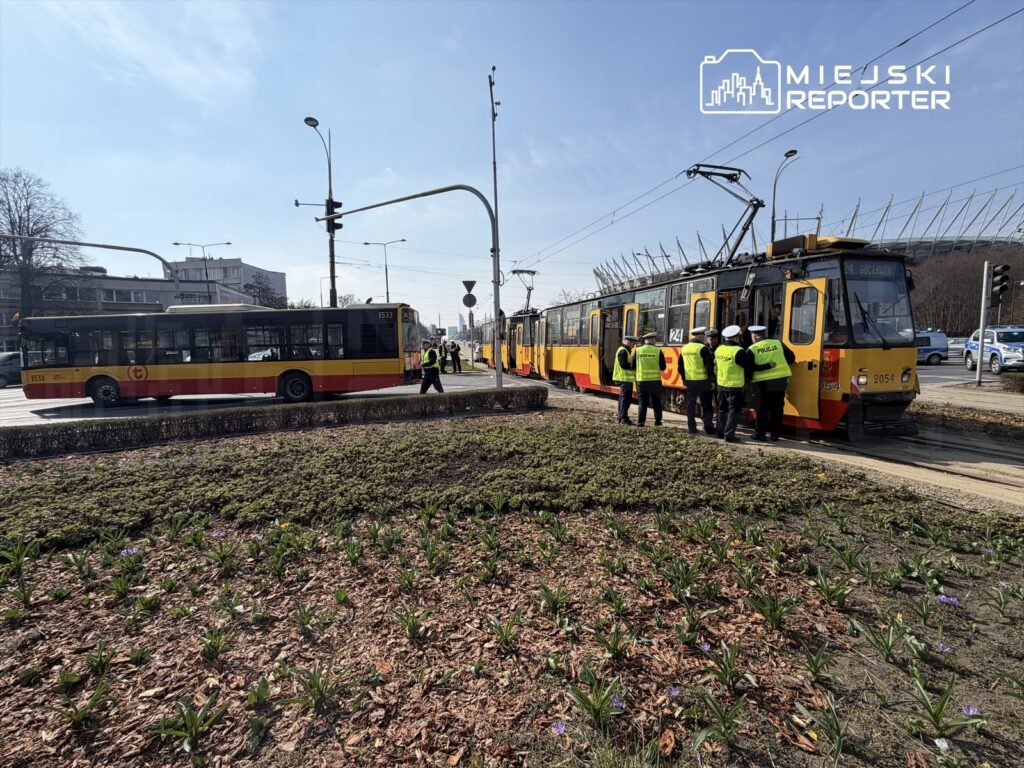 Grupa pracowników w kamizelkach odblaskowych sprawdza tramwaj na przystanku obok autobusu w miejskim otoczeniu.