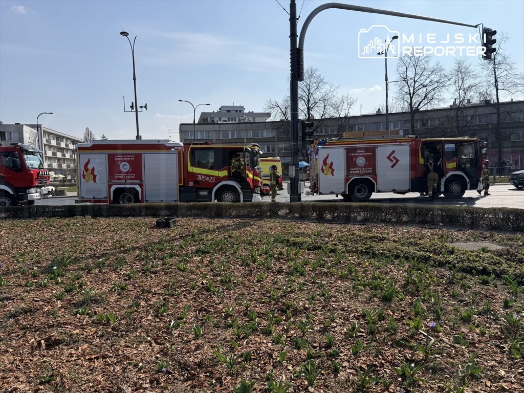 Wozy strażackie zaparkowane w pobliżu skrzyżowania, obok nowoczesnych budynków mieszkalnych, wiosenne rośliny na pierwszym planie.