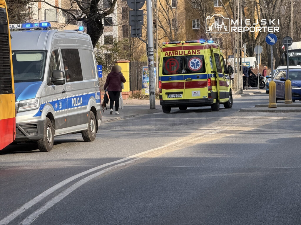 Policyjny van z niebieskimi sygnałami świetlnymi zatrzymany na ulicy, obok przejeżdżająca karetka.