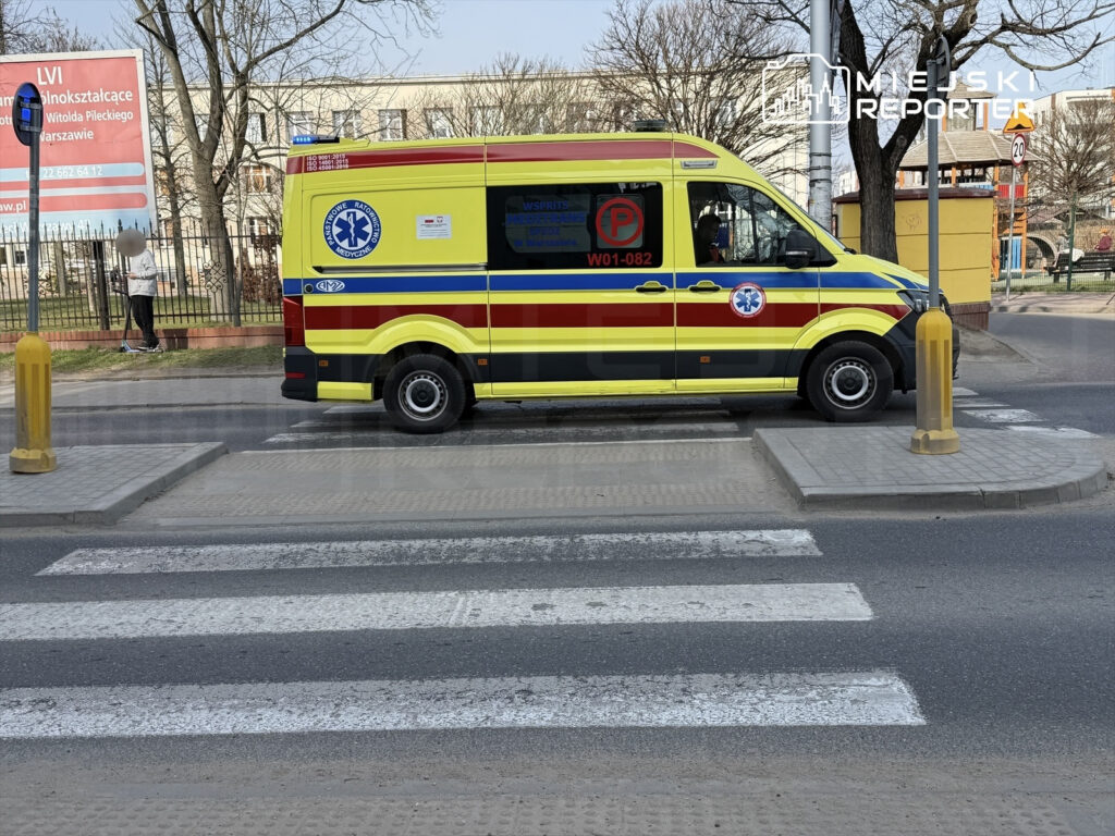 Karetka pogotowia w żółto-czerwonym kolorze przejeżdża przez przejście dla pieszych w miejskim otoczeniu.