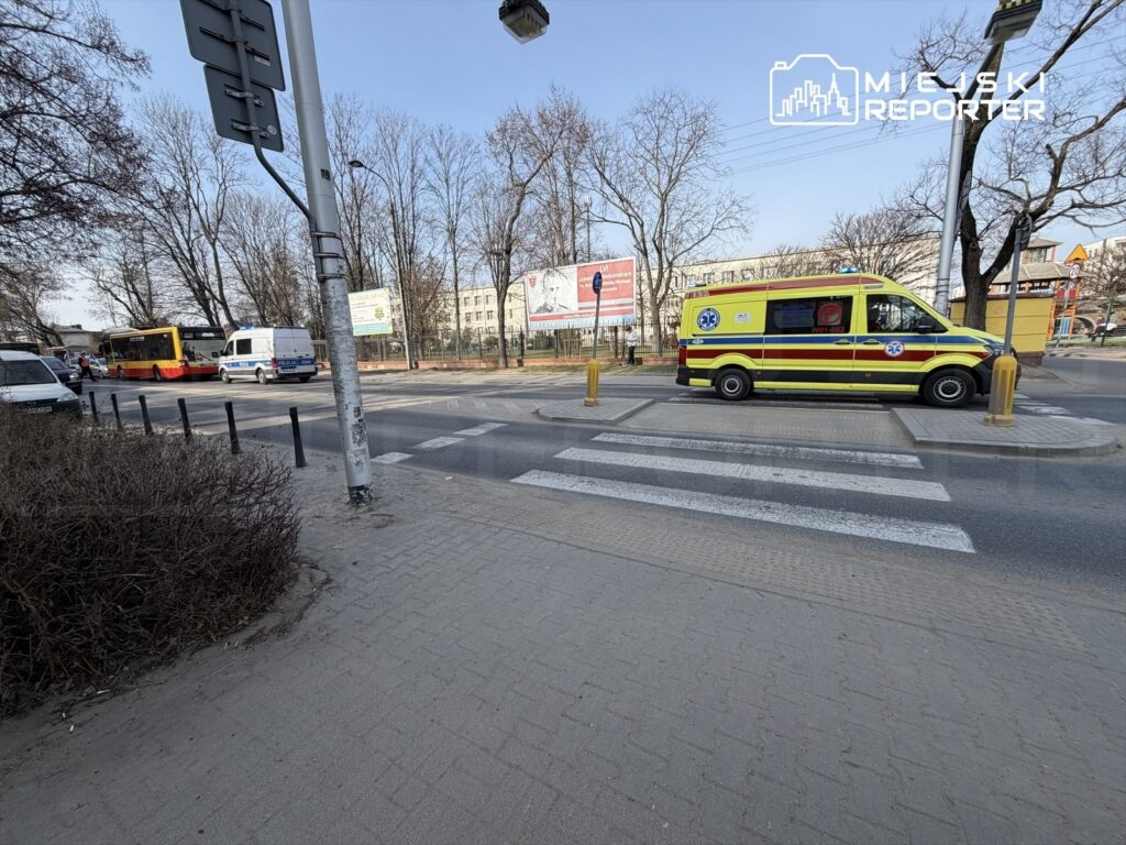 Karetka pogotowia przejeżdża przez przejście dla pieszych obok ruchliwej ulicy z samochodami i drzewami w tle.