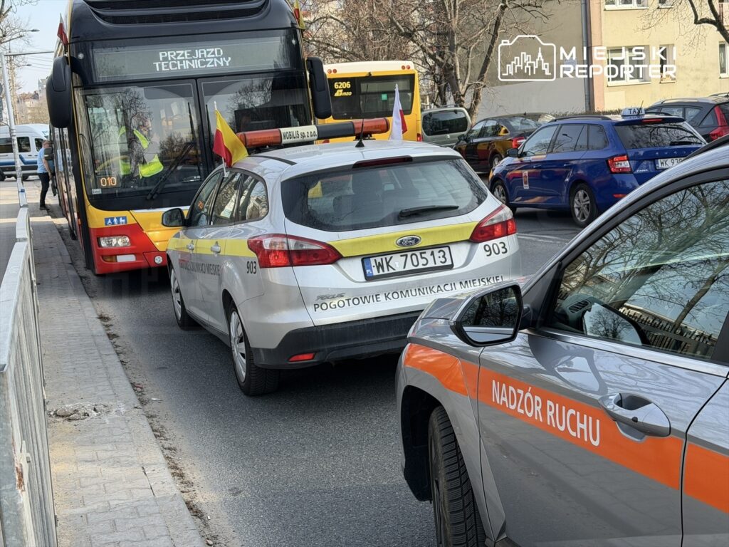 Przed autobusami z flagami, samochód Pogotowia Komunikacji Miejskiej zatrzymany obok pojazdu Nadzoru Ruchu.