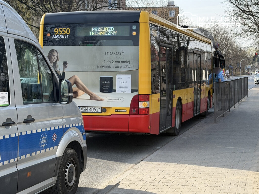 Autobus miejski z reklamą kosmetyków zatrzymany na przystanku, obok stoi radiowóz policyjny.