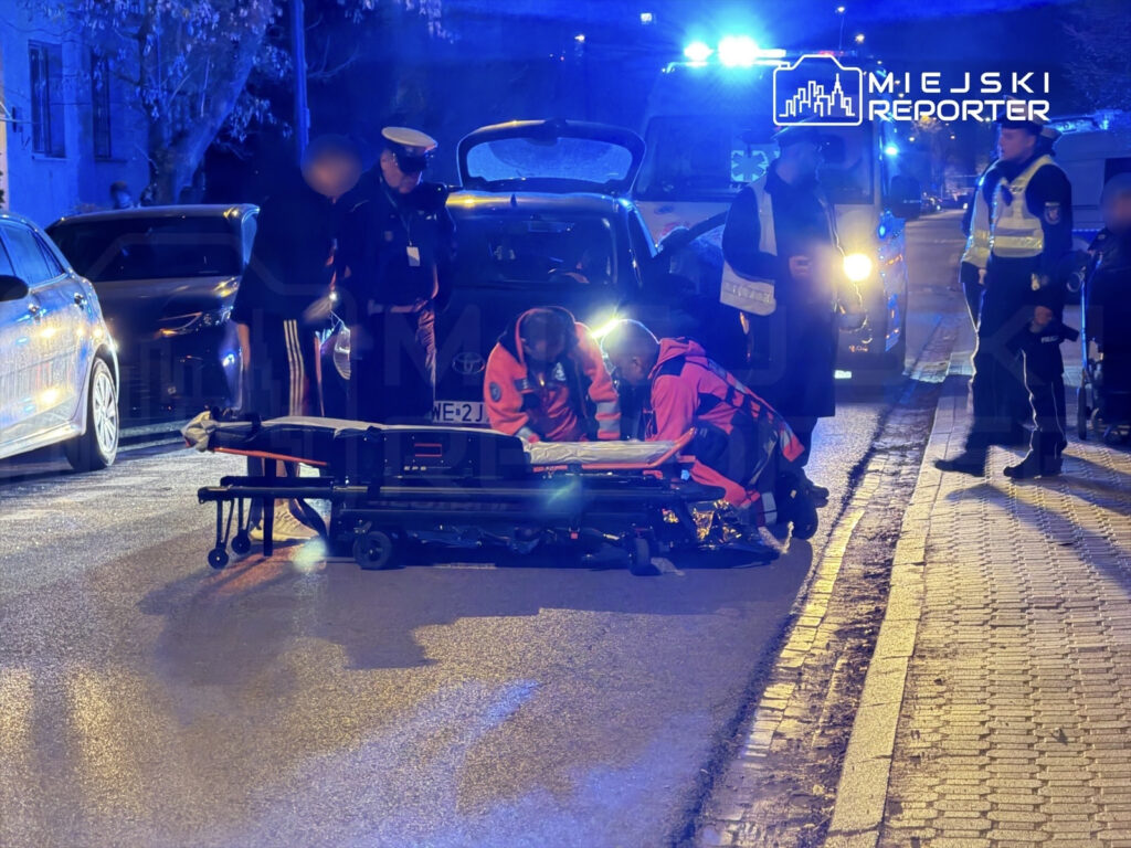 Ratownicy medyczni pracują nad osobą leżącą na noszach na oświetlonej ulicy, w pobliżu pojazdów i funkcjonariuszy policji.