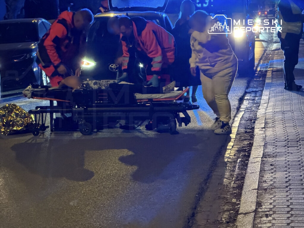 Ratownicy medyczni udzielają pomocy osobie leżącej na noszach na ulicy, w pobliżu zaparkowanych samochodów.
