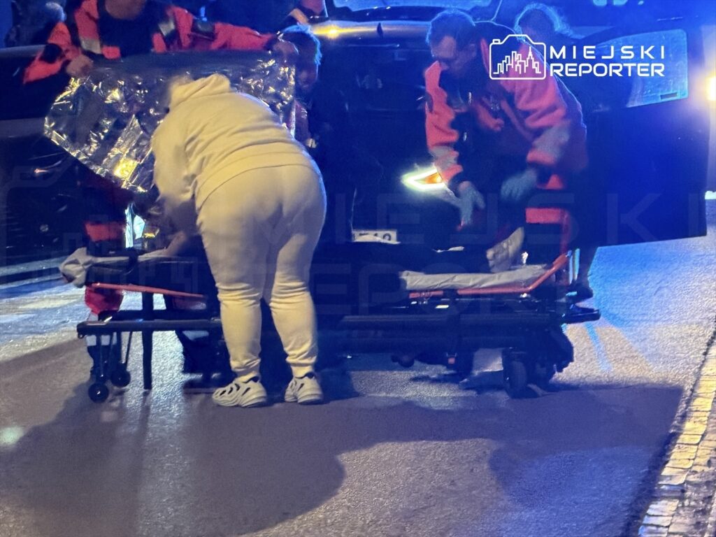 Ratownicy medyczni udzielają pomocy poszkodowanemu na noszach w nocy, otoczeni przez pojazdy i policyjne światła.