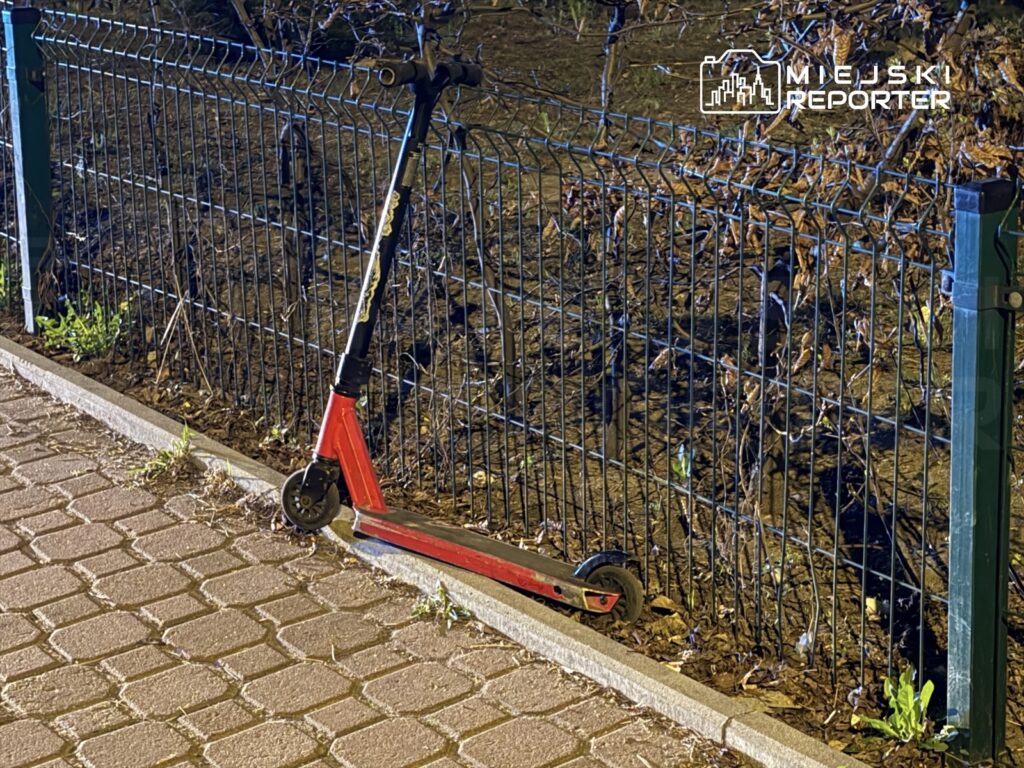 Czerwona hulajnoga oparta o ogrodzenie w nocy, na chodniku z kostki brukowej, w tle widoczne uschnięte rośliny.