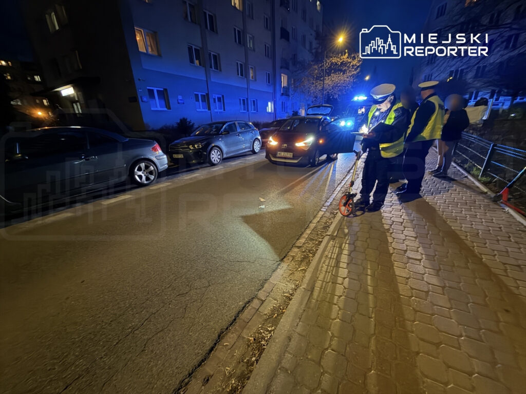 Funkcjonariusze policji mierzą odległość na drodze w nocy, otoczeni zaparkowanymi samochodami.