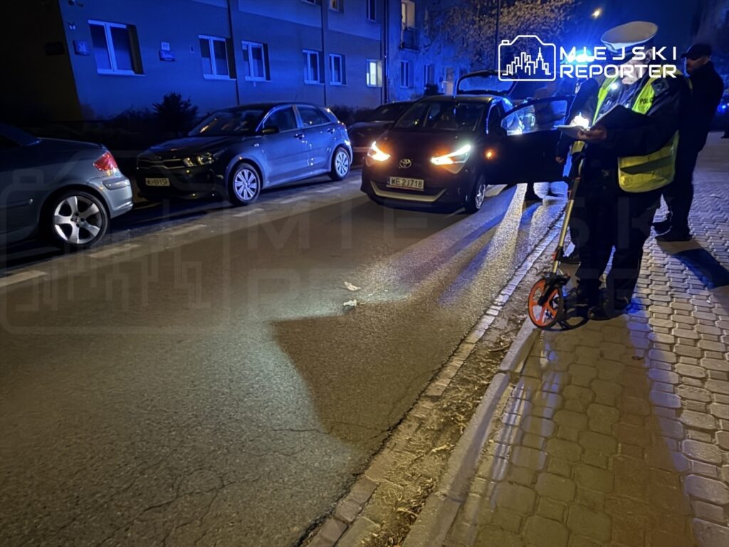 Policjant w kamizelce odblaskowej mierzy odległość na oświetlonej ulicy, obok zaparkowanych samochodów.