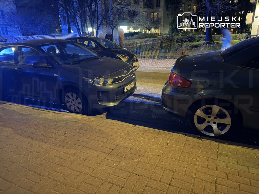 Dwa zaparkowane samochody, jeden szary, drugi czarny, stoją blisko siebie na oświetlonym chodniku.