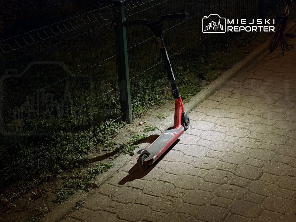 Czerwona hulajnoga elektryczna stoi na oświetlonym chodniku obok metalowego ogrodzenia w nocy.