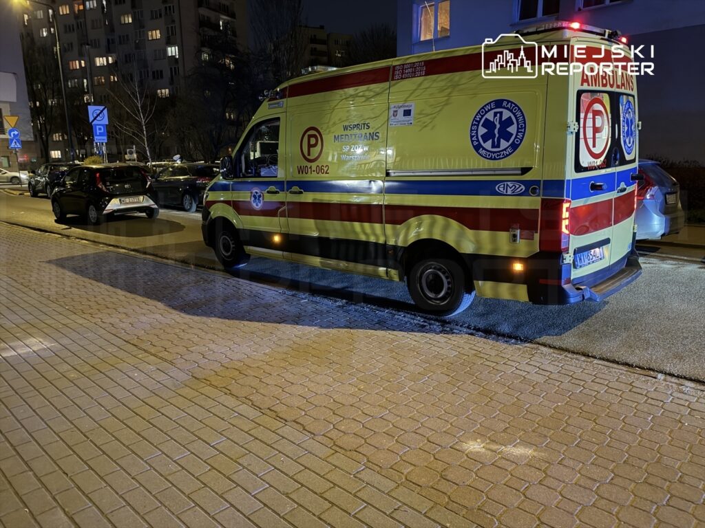 Karetką pogotowia zaparkowaną wzdłuż ulicy, oświetloną przez latarnie, obok zaparkowanych samochodów.
